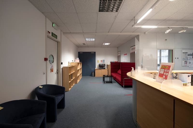 The image shows an office space with a reception area, shelves, a red sofa, and various office equipment and furniture. The space appears to be well-lit with ceiling lights and has a professional and functional layout.