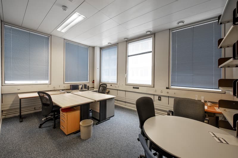 large empty office room, white walls, fluorescent ceiling lights, three windows with blinds, round and rectangular desks, file cabinets, and chairs