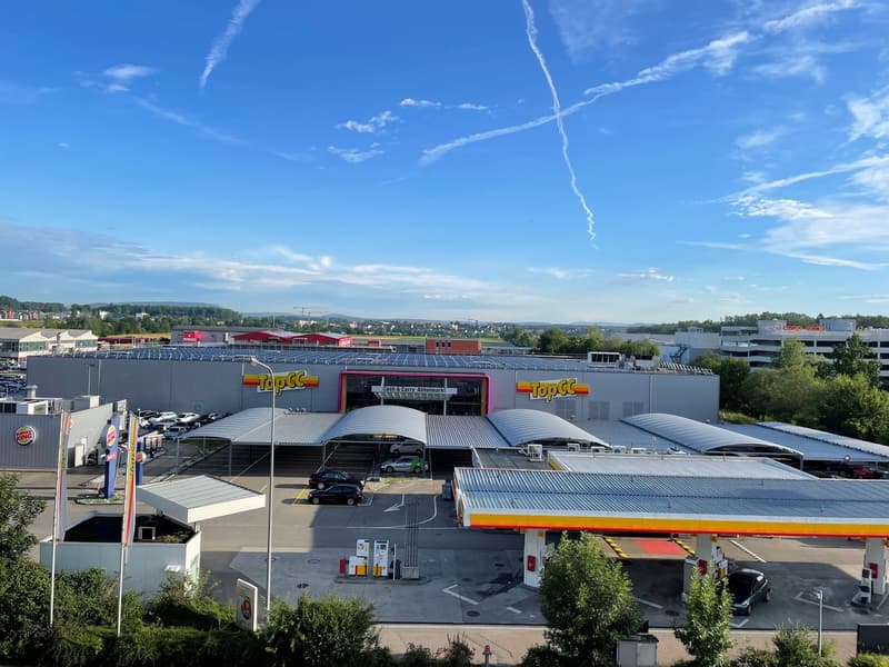 Retail building with a gas station, multiple cars parked, solar panels on the roof, a McDonald's franchise in front, and a surrounding urban area with trees and mountains in the background.