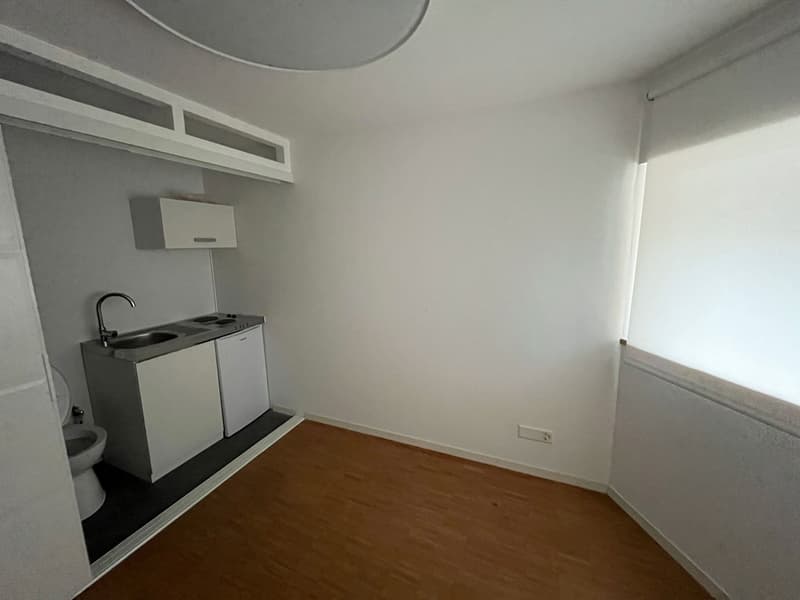 white walls, wooden floor, open kitchen with sink, electric stove, small white cabinets, toilet
