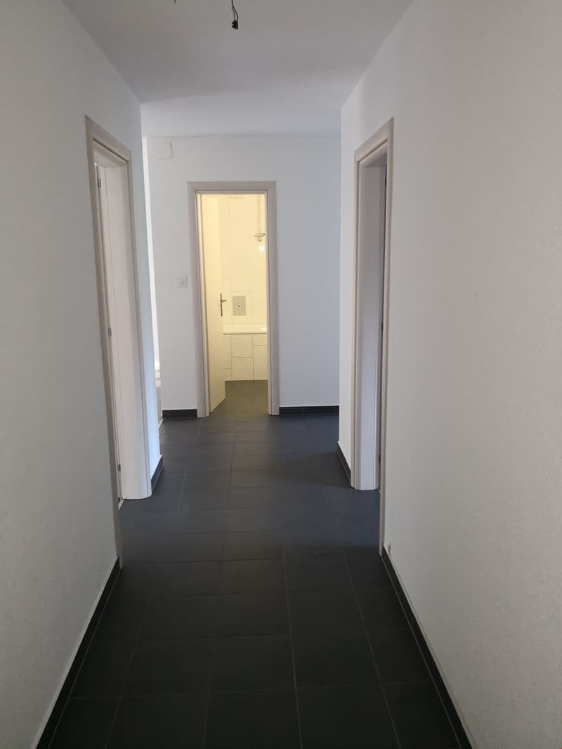 The image shows a hallway with white walls and dark tile flooring. There are two doorways visible, one of which appears to be the entrance to another room. The hallway has a clean, minimalist design.