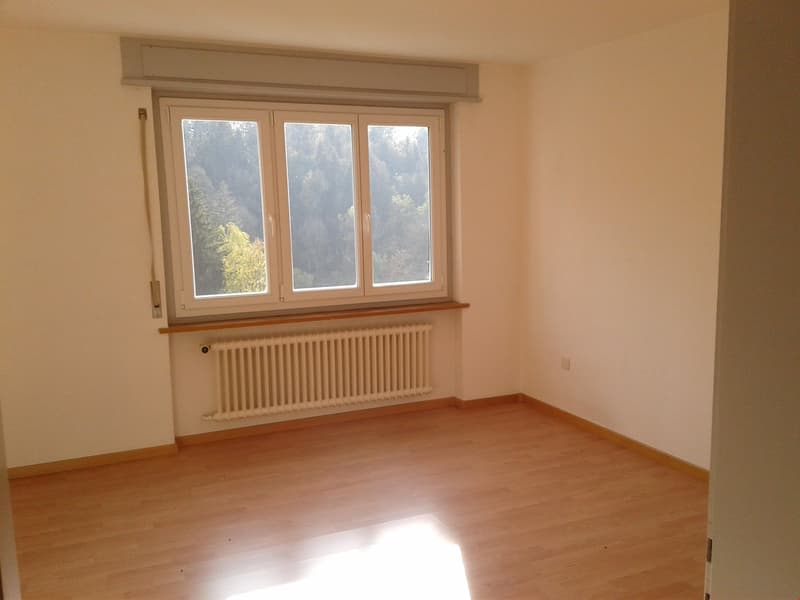 The image shows a spacious bedroom with large windows overlooking a forested area. The room has hardwood floors and a radiator along the wall. The room appears to be empty, with no furniture or decor visible.