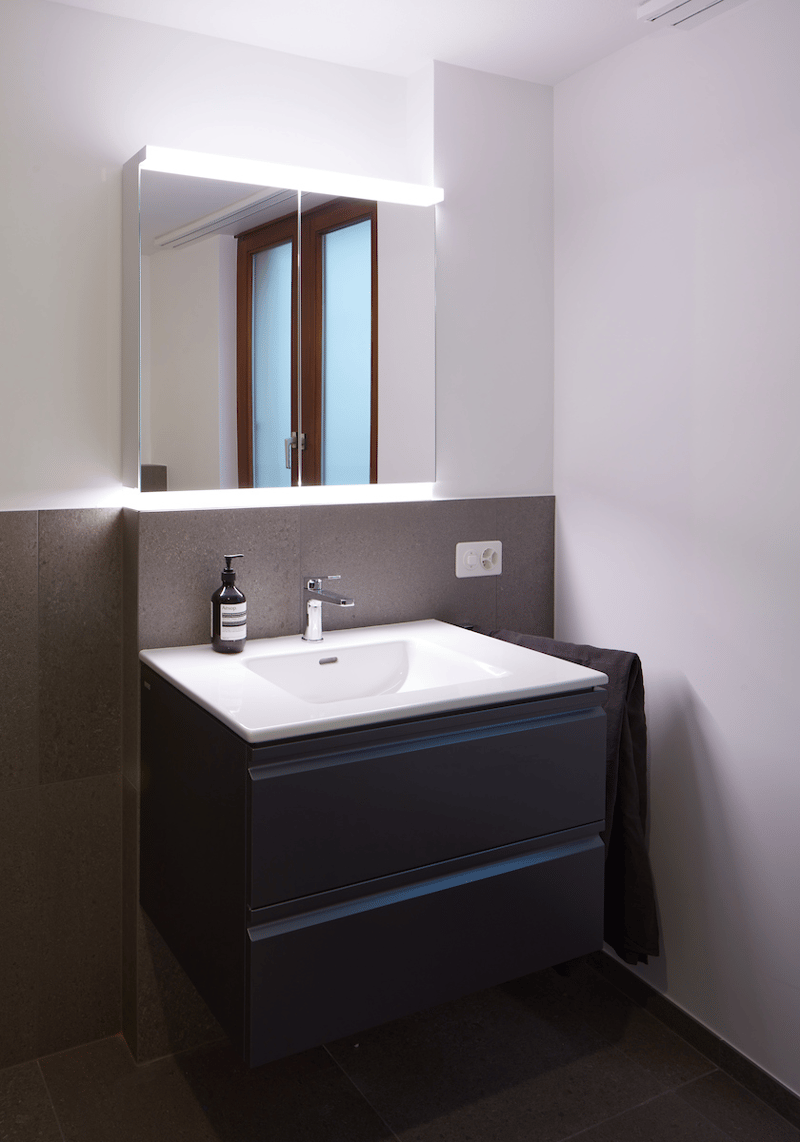 Bathroom: white tiled floor, white sink, large mirror, dark grey cabinetry, built-in towel rack, power outlet, wooden window.