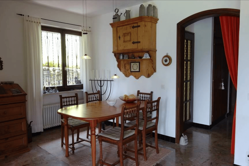 Wooden table, chairs, kitchen cabinets, radiators, hanging shelves, decorative items, chandeliers, bowl with fruits, door