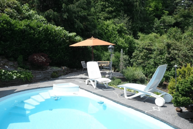 Swimming pool, white lounge chairs, orange umbrella, gravel, table and chairs, plants and trees, mountain in background