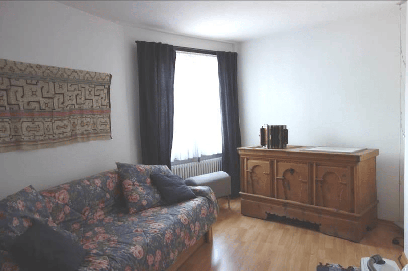 a wooden floored living room with a floral couch, a wooden cabinet, and a window with curtains