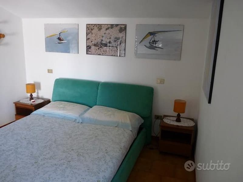 bedroom with a bed, two nightstands, and a wall with three paintings. The bed is made with two white pillows and a floral blanket. Two nightstands on either side of the bed have lamps and a power outlet each.
