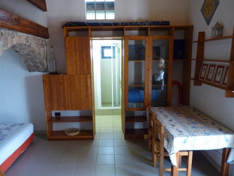 A room with wooden cabinets, a table, and a bed. There is a bed placed against the wall, a wooden shelf is on the left side, and a dining table is placed near the wall. The cabinet is mounted on the wall, and the person is standing near the cabinet. There
