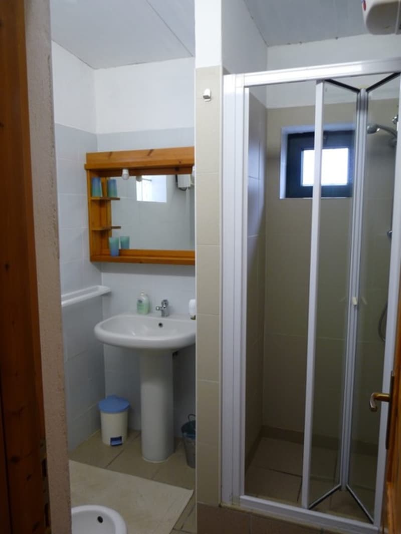 white tiled walls, wooden shelf with cups, sink with faucet, walk-in shower, white bidet, trash bin