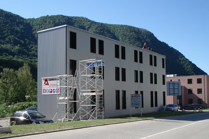 3-story gray industrial or commercial building with multiple windows, scaffolding on the side, and parked cars in the foreground. The building is situated in a mountainous, forested area.