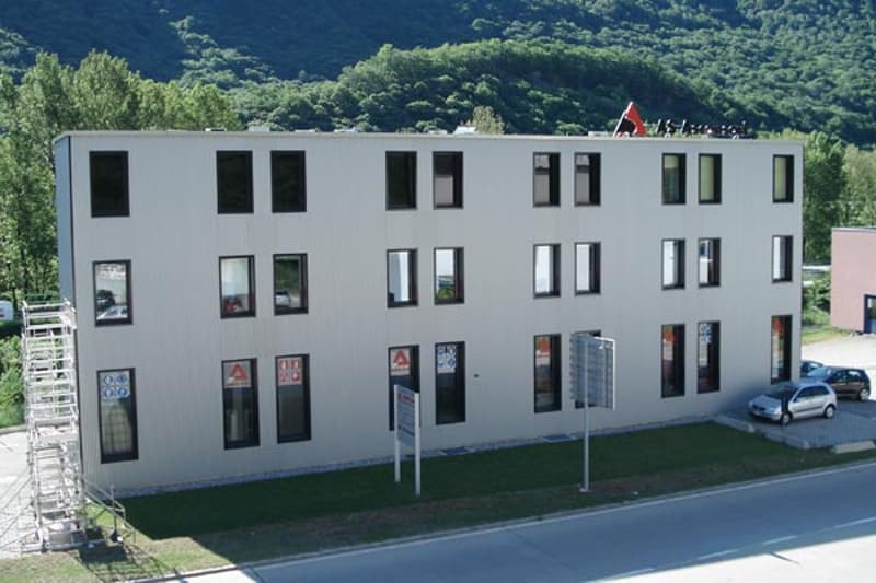 3-story commercial or office building with a gray exterior, multiple windows, and a parking area in front.