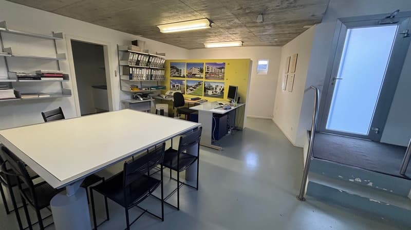An empty office with tables, chairs, monitors, and shelves. Includes an open door to another room, a ceiling with multiple lamps, and stairs leading up to a higher floor.