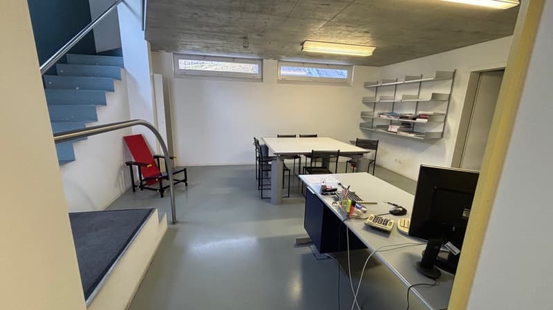 Empty room with white walls, tables, chairs, computer monitors, keyboard, shelves with books, grey carpet, staircases, windows, ceiling lights