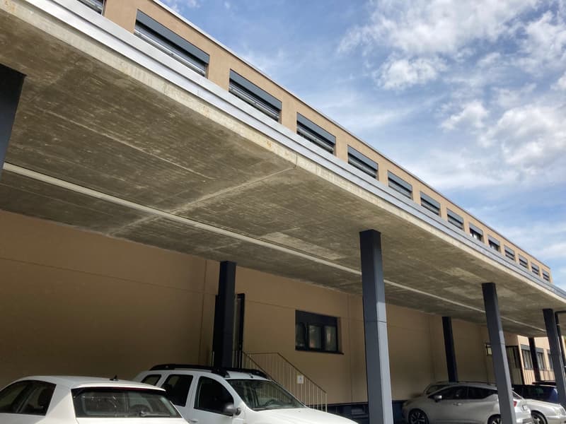 Building with a covered parking area, concrete ceiling, beige walls, multiple windows, and cars parked underneath.