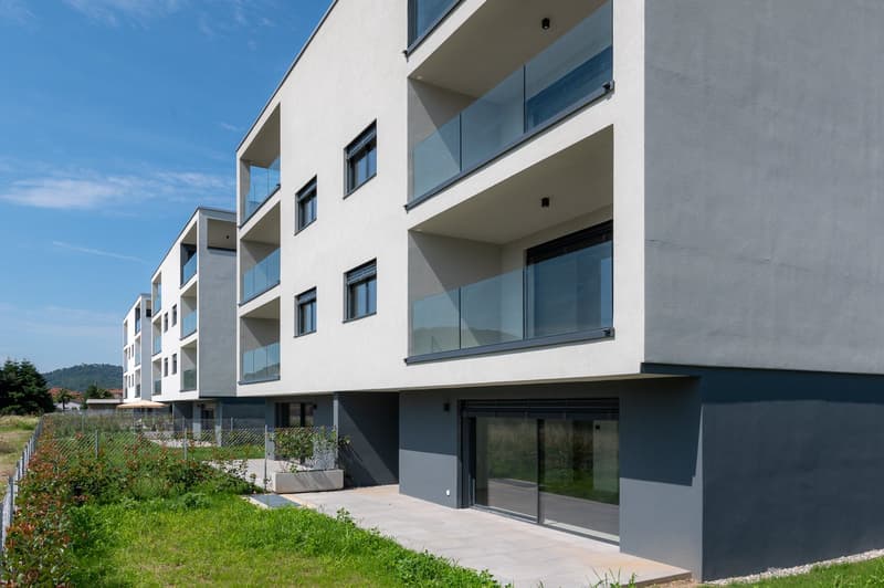 Multi-story apartment building with modern, minimalist design. The building has large windows and glass balconies on each floor. The exterior is a combination of white and gray colors. There is a small garden area in front of the building.
