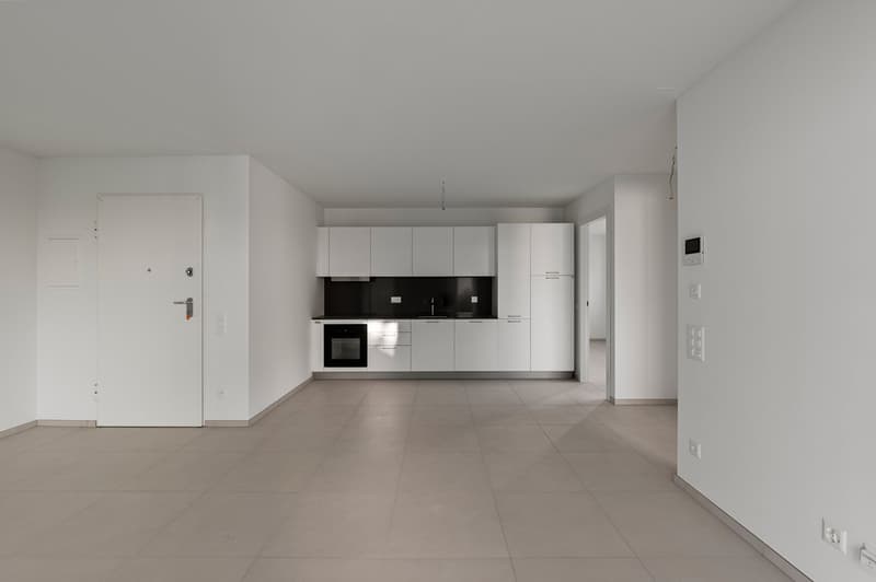 The image shows a modern, open-plan apartment with a kitchen and living area. The apartment has white walls, gray tile flooring, and a minimalist white kitchen with built-in appliances. The space appears to be newly renovated and has a clean, minimalist a