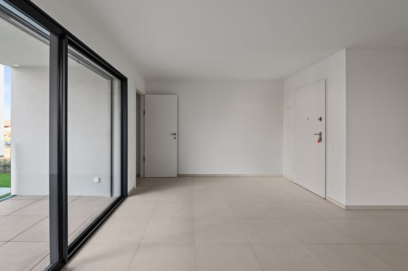 Spacious room with large sliding glass doors, white walls, and light-colored tile flooring. The room appears to be empty, with a white door visible on the right side.