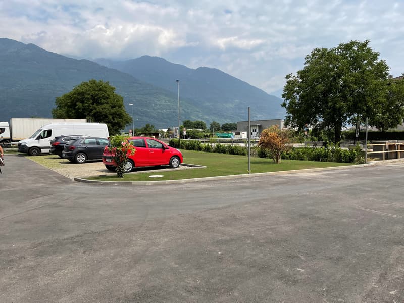 Empty parking lot with several cars parked, surrounded by trees and bushes, mountains in the background