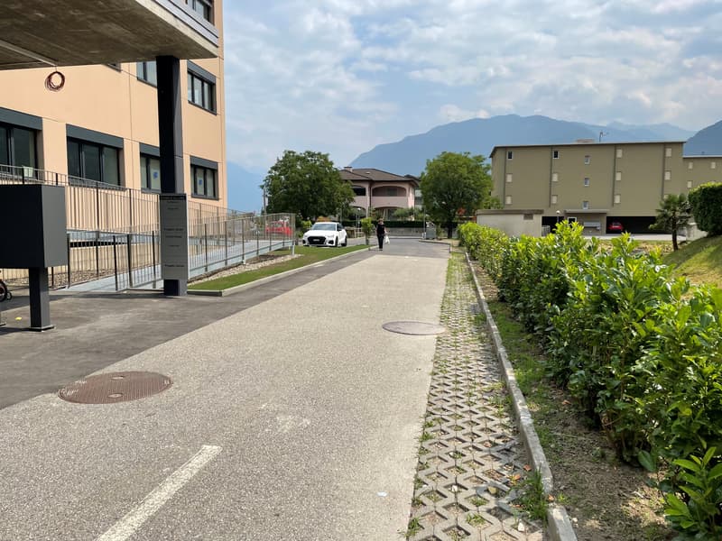 A paved road with a sidewalk, a building with a metal fence, a parked white car, a person walking, and a mountain in the distance.