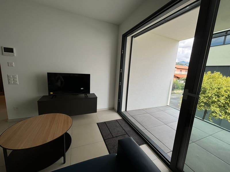 Living room: couch, wooden coffee table, black TV cabinet, flat screen TV, sliding glass door to terrace, beige tiles.