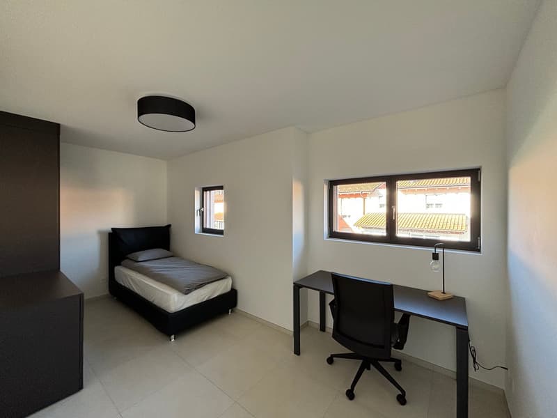 The image shows a bedroom with a single bed, a desk with a chair, and large windows providing natural light. The room has a minimalist and modern design with white walls and tile flooring.