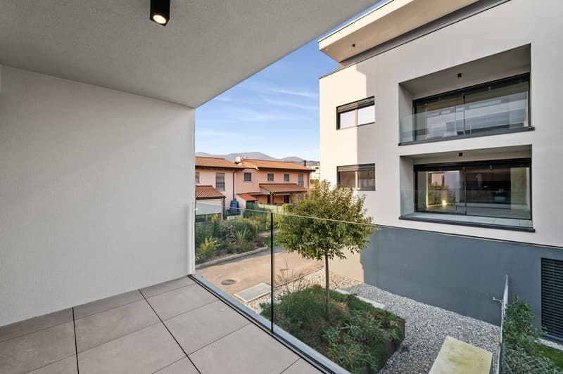 The image shows a modern apartment unit with a balcony or terrace overlooking a landscaped garden area. The apartment has large windows and sliding glass doors that provide access to the outdoor space. The building appears to be part of a residential comp