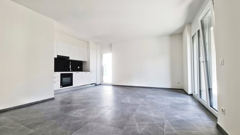 Open-plan living room and kitchen with white walls, large windows, grey tile flooring, and modern kitchen appliances including a dishwasher.