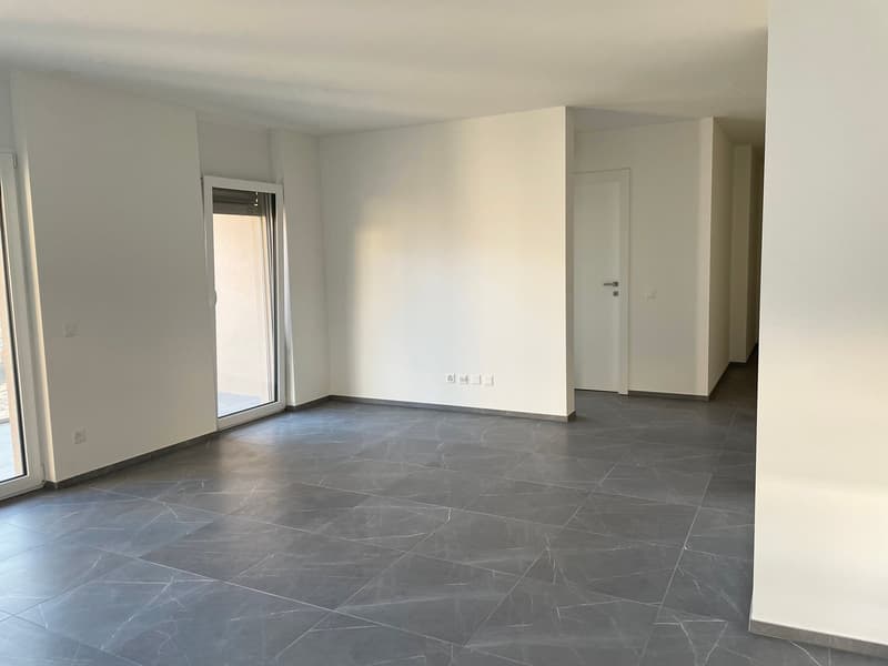 This image shows a spacious room with white walls and a large window. The floor is covered with gray marble-like tiles, creating a sleek and modern look. The room appears to be empty, with no furniture or decor visible.