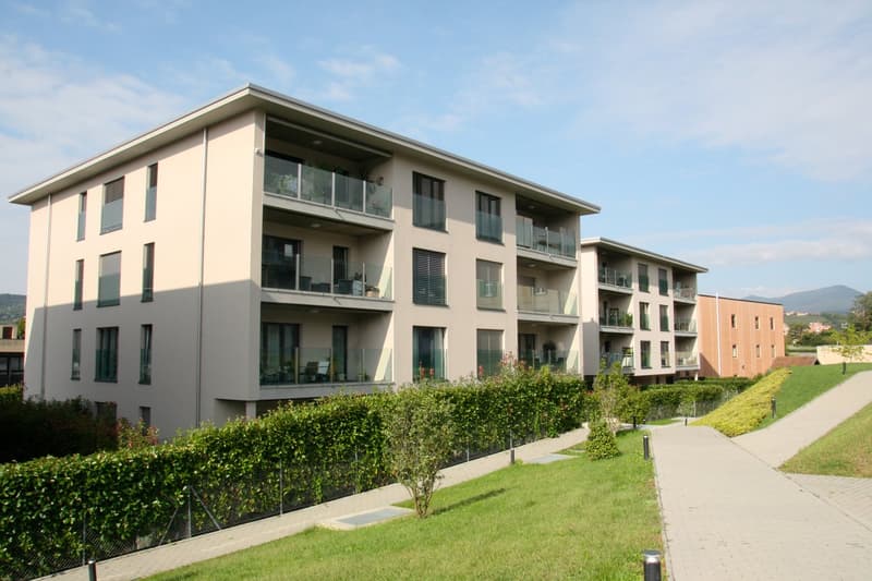 Modern 3-story apartment complex, white exterior, glass balconies, hedged garden, paved walkway, green lawn, plants, and shrubs.