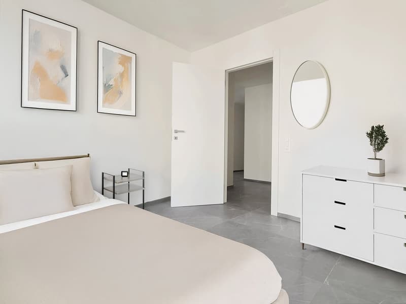 Bedroom with a large double bed, pillows, nightstand with a small table, white cabinet, decorative mirror, wall art paintings, open door leading to another room.