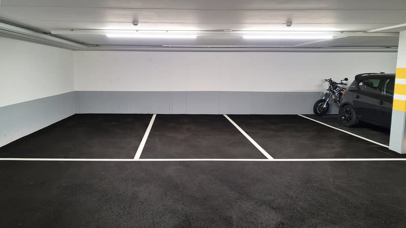 Spacious underground garage with multiple parking spaces, concrete floor, and white walls. A motorcycle is parked in one of the spaces.