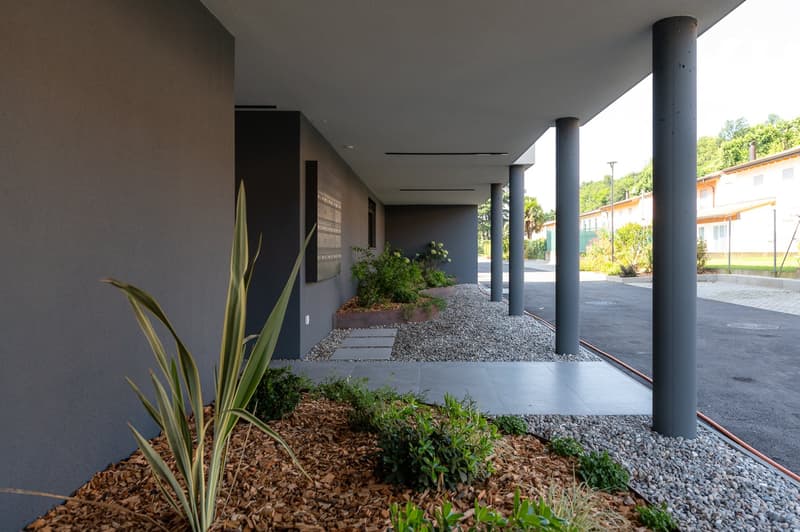 A building with a modern grey exterior wall, gravel walkway, planted flower beds, and two vertical poles