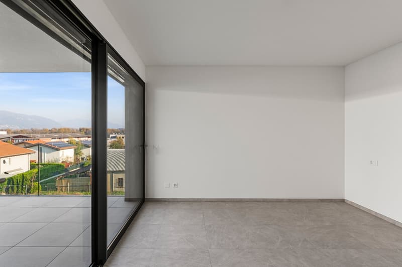Spacious room with large windows, providing a view of the surrounding buildings and landscape. The room has white walls and grey tile flooring.