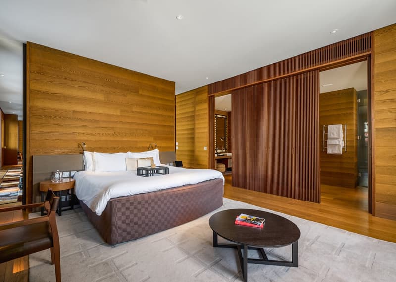 A bedroom with wooden walls, a large bed, a wooden coffee table, a chair, and a side table with a bottle on it.