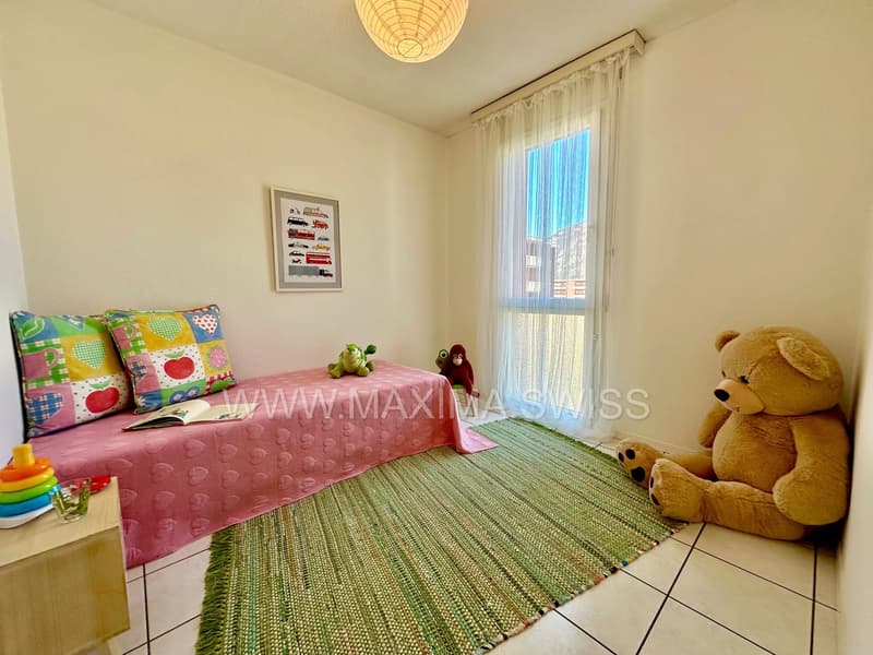 Small bedroom with a pink bed, colorful pillows, white tiled floor, rug, stuffed animals, books, and decorations