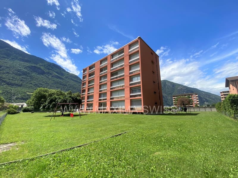 Apartment building with multiple floors, large windows, green lawn, trees, mountains in the background