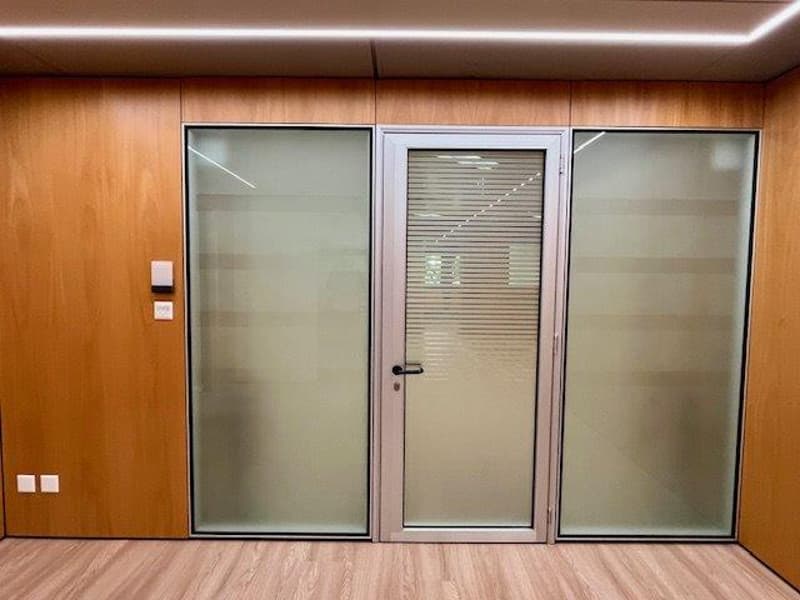 The image shows a room with wooden walls and a glass door in the center. The glass door appears to have a shutter or blinds inside, suggesting it may lead to a balcony or terrace. The room has a hardwood floor and appears to be a modern, minimalist design