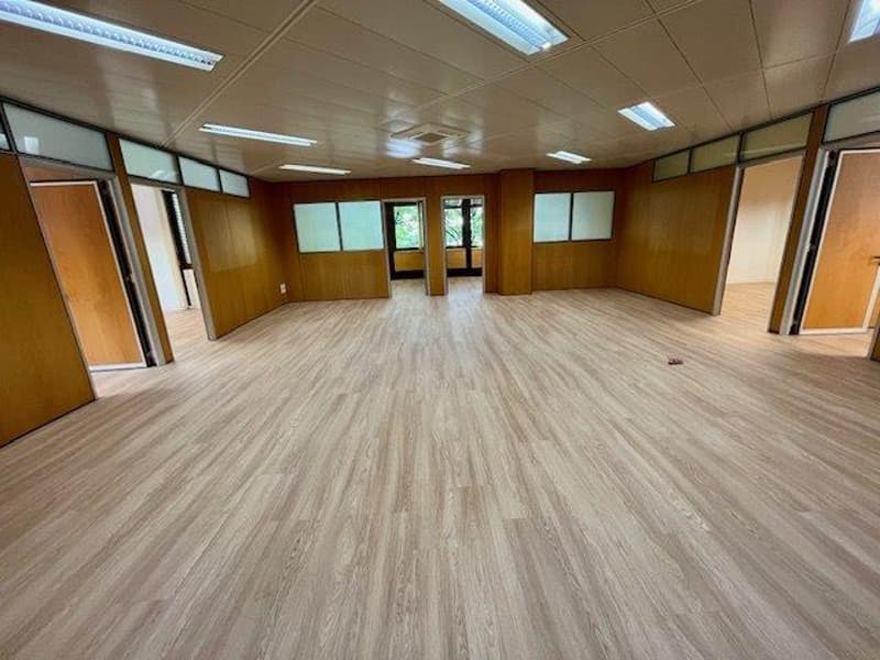 The image shows a large, open office space with a wooden floor and walls. The room has multiple windows and lighting fixtures, and appears to be a commercial or industrial space that could be used for various purposes such as an office, meeting room, or e