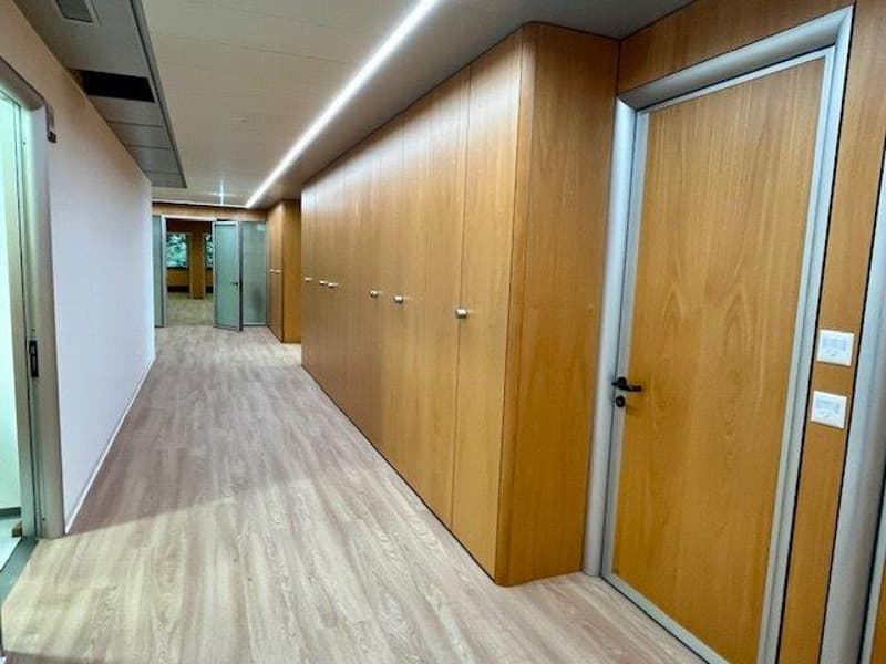 The image shows a long hallway with wooden paneled walls and doors on both sides. The floor is covered with a light-colored laminate or vinyl flooring. The hallway is well-lit with recessed lighting fixtures in the ceiling.