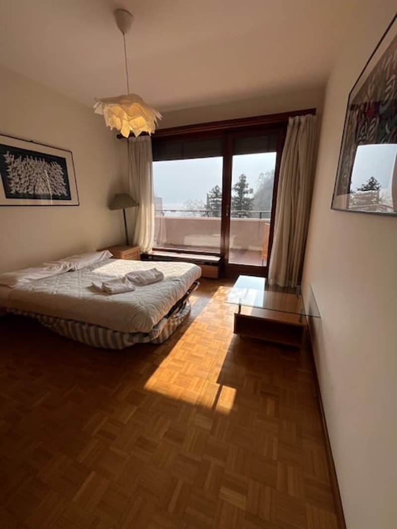 A bedroom with a bed, a glass table, a lamp, and large windows leading to a balcony with a view of the surroundings.
