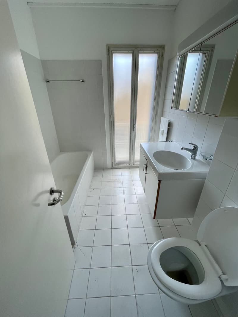 white tiled floor, white walls, bathtub, white sink with silver faucet, large mirror above sink, toilet, silver radiator