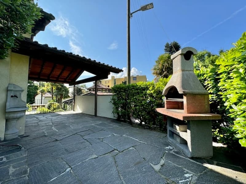 Outdoor area with stone paving, brick oven, covered area with wooden roof, greenery, plants, and trees