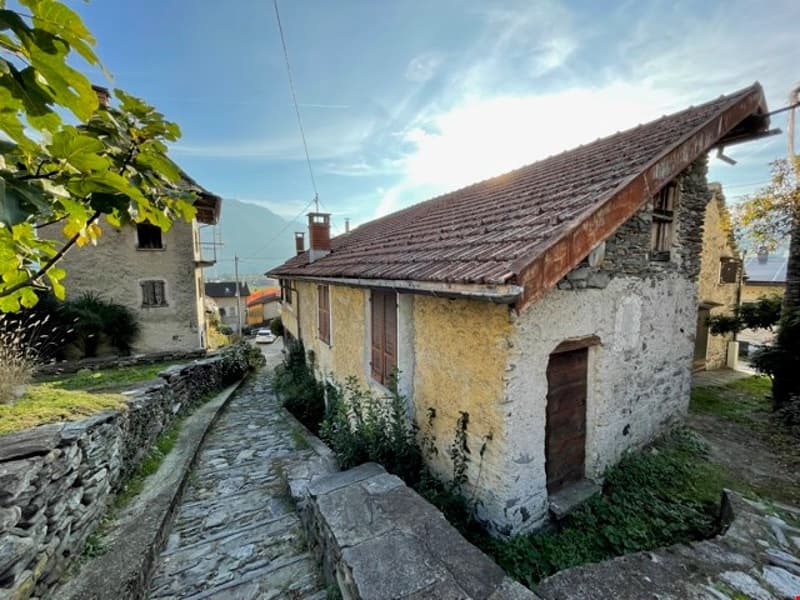 an old stone house, with a brown roof, a narrow street with a slope, the road with stones, the house on the left side, a house in the distance