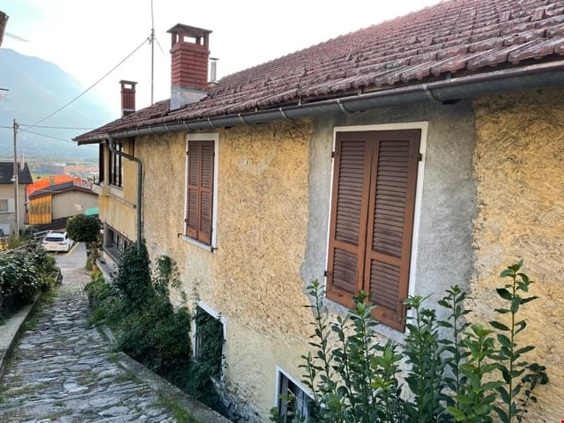 2 story house, beige wall, brown roof, brick chimney, balcony, 2 windows with brown shutters, green plants in front