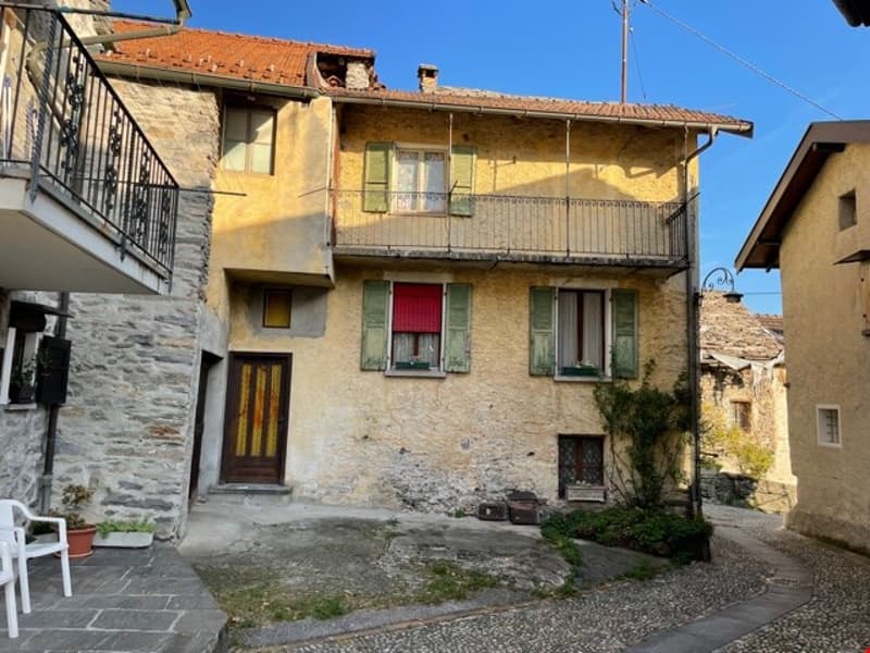 Yellow stone building, red roof, brown wooden door, balcony, green shutters, plants, narrow street, nearby houses