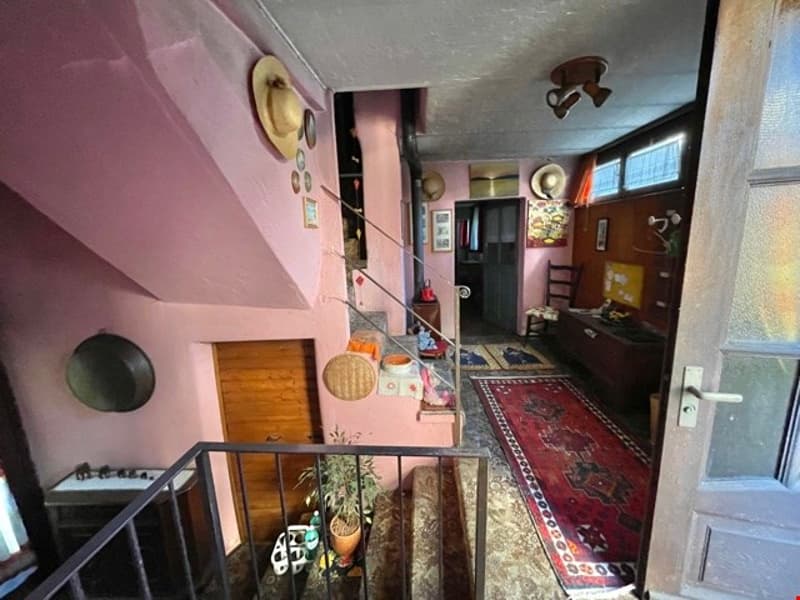 Pink walls, white tiled floor, a red rug, a balcony with a metal railing, a staircase with handrails, a small desk with objects, and a hat hanging on the wall