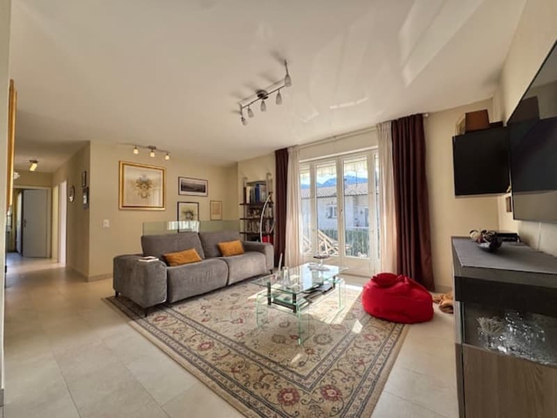 Spacious living room with large windows, gray sofa, glass coffee table, and area rug. The room has a balcony or terrace visible through the windows.