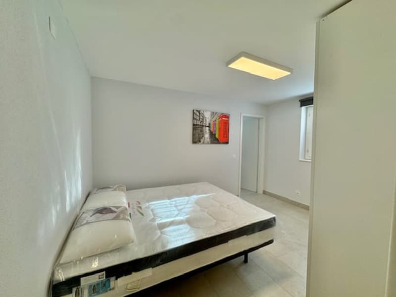 Spacious bedroom with white walls, hardwood flooring, and a large bed with a mattress. There is a framed artwork on the wall and a ceiling light fixture providing illumination.