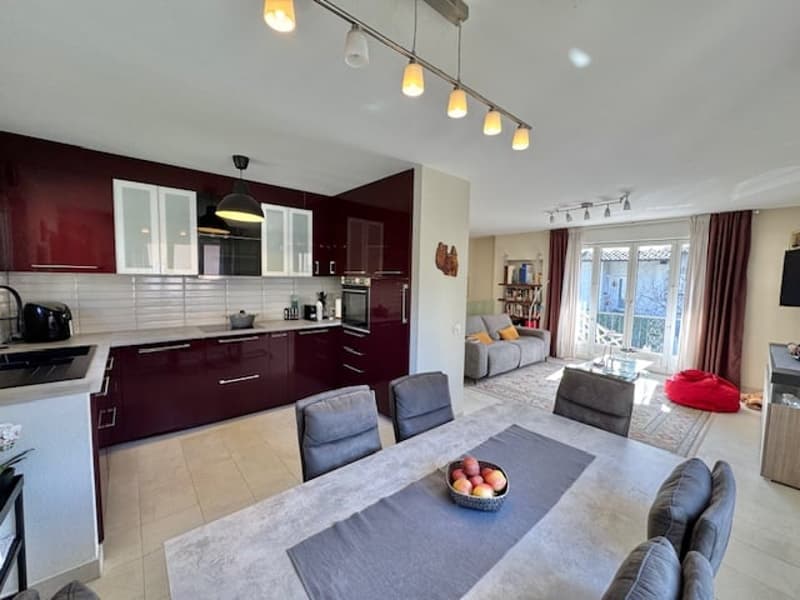 The image shows an open-plan living room and kitchen area. The kitchen features burgundy cabinets and a modern design. The living room has a gray sofa and chairs, and a glass door leading to a balcony or terrace. The space is well-lit with track lighting