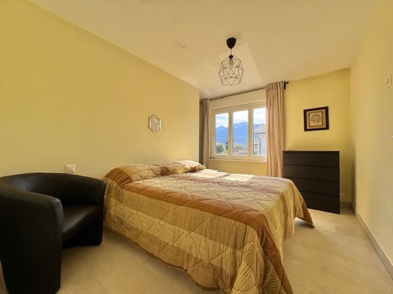 Spacious bedroom with large windows providing a scenic view, beige walls, hardwood flooring, a king-size bed with a patterned comforter, and a black leather chair.